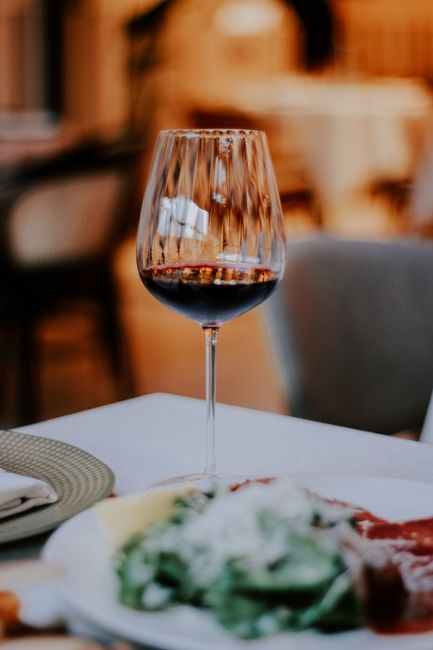 Close-up of a wine glass tipped over, spilling red wine onto a white surface in a clean, well-lit room. The glass, made of clear glass with a thin stem and rounded bowl, is partially filled with red w