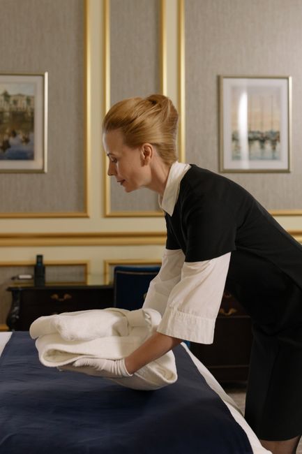 A female cleaning professional with blonde hair, smiling and engaged in a conversation, standing in front of a window with sheer white curtains that allow natural light to fill the room. She is wearin