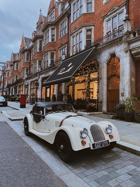 An exterior view of a red-brick townhouse with large sash windows, ornate stone archways, and a black awning with gold lettering, located on a paved street in Mayfair. A vintage cream-colored converti
