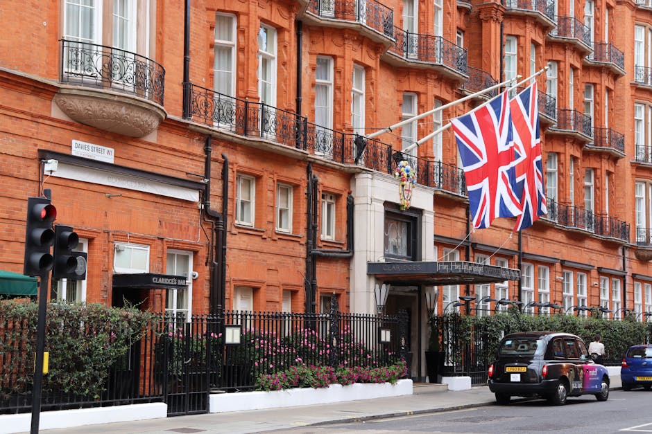 An exterior view of a red-brick townhouse with large sash windows, ornate stone archways, and a black awning with gold lettering, located on a paved street in Mayfair. A vintage cream-colored converti
