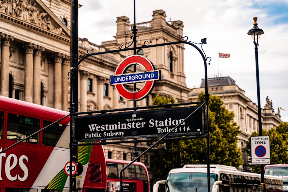 Close-up view of the Westminster Station sign, featuring the classic London Underground red circle and blue bar, mounted on an ornate black iron frame. Behind the sign, a historic stone building with 